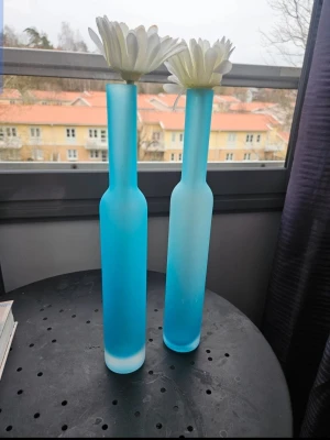 9 höga blå glasvaser - Säljer 9 stilrena, höga vaser i frostat blått glas med smal hals och cylinderformad kropp. Perfekta för enstaka blommor eller som dekorativa inredningsdetaljer. Ger ett modernt och fräscht intryck till rummet. Användes under en konfirmationsmottagning. Skickar med blomman i vaserna. Kan även hämta sig Partille