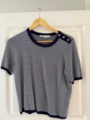 Randig topp från Zara med knappar - Snygg kortärmad topp från Zara med marinblå och vita ränder. Rund halsringning och dekorativa knappar på ena axeln. Perfekt till jeans eller kjol för en fräsch look.