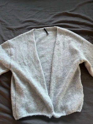 Ljusgrå öppen stickad kofta - Mysig ljusgrå stickad kofta med öppen front och lång ärm. Koftan har en avslappnad passform och är tillverkad i ett mjukt, fluffigt material som känns skönt mot huden. Perfekt att slänga på sig när det är lite kyligt ute.