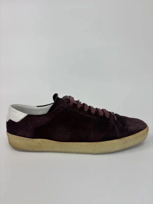 Vinröda Saint Laurent sneakers i mocka - Snygga vinröda sneakers i mocka med matchande vinröda skosnören och vit detalj på hälen. Yttersulan är beige och ger en retrovibe. Perfekta för dig som gillar stilrena och klassiska sneakers med en twist.