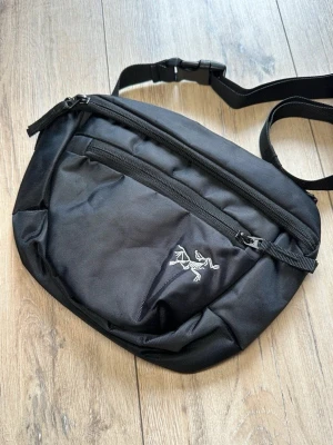 Svart väska från Arc'teryx - Snygg svart axelväska från Arc'teryx med justerbar rem och flera dragkedjefack. Väskan har en stilren design med Arc'teryx-loggan broderad framtill och är tillverkad i slitstarkt material. Perfekt för att bära dina viktigaste grejer på ett smidigt sätt.