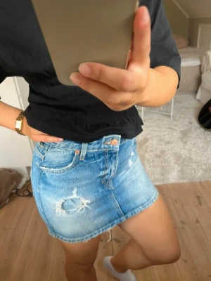 Blå jeanskjol med slitningar - Snygg kort jeanskjol i ljusblå denim med slitna detaljer och klassisk femficksdesign. Kjolen har en rak passform och råa kanter nertill för en trendig look. 