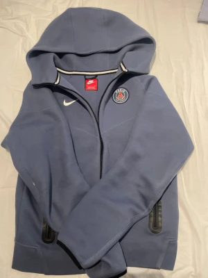 Blå Nike PSG hoodie med dragkedja - Snygg blå hoodie från Nike med PSG-logga på bröstet och vit Nike Swoosh. Hoodien har dragkedja framtill, två fickor med dragkedja och stor huva. Tillverkad i en mjuk bomull som känns skön mot huden. Perfekt för dig som gillar sportig stil. Det är en väldigt skön och snygg hoodie
