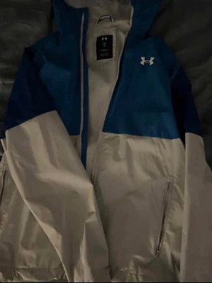 vindjacka Under Armour - Snygg tvåfärgad vindjacka från Under Armour i blått och vitt. Jackan har huva, dragkedja framtill och två sidofickor med dragkedja. Tillverkad i lätt och tåligt material som står emot vind. Perfekt för dig som vill ha en sportig look.