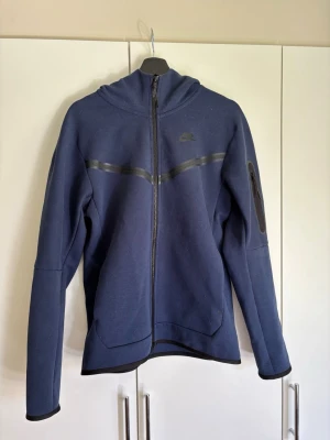 Mörkblå hoodie med dragkedja - Snygg mörkblå hoodie från Nike  med hel dragkedja framtill och huva. Har en diskret logga på bröstet och en svart detalj över bröstet. Ficka med dragkedja på ena ärmen. Perfekt för en sportig och avslappnad stil.