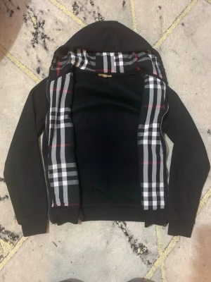 Svart hoodie från Burberry med rutigt foder - Snygg svart hoodie från Burberry med dragkedja och klassiskt rutigt foder i huvan och längs insidan. Hoodien har långa ärmar, ribbade muddar och en skön passform. Perfekt för dig som gillar stilrena plagg med en twist.
