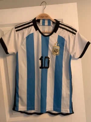 Argentina Messi fotbollströja och shorts  - Säljer en klassisk Argentina fotbollströja med matchande shorts från Adidas med blå och vita ränder, svarta detaljer och nummer 10. Messi står på ryggen. Tröjan har korta ärmar, rund hals och AFA-emblem på bröstet. Perfekt för fotbollsfans! Storlek xs