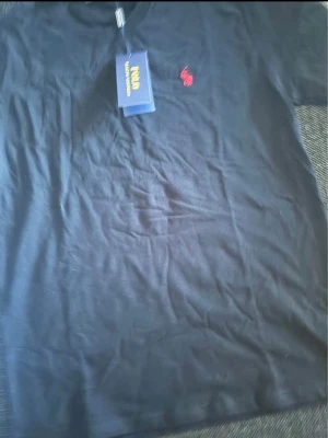 Svart t-shirt från Polo Ralph Lauren - Klassisk svart t-shirt från Polo Ralph Lauren med röd broderad logga på bröstet. T-shirten har korta ärmar och rund halsringning. Tillverkad i mjuk bomull som känns skön mot huden. Perfekt basplagg till garderoben.