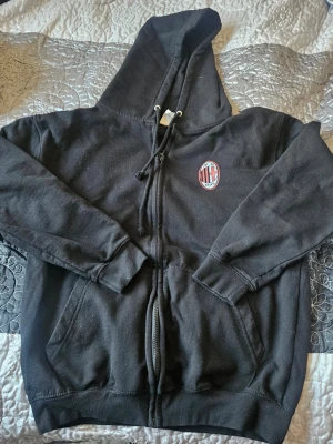 Svart AC Milan hoodie med dragkedja - Svart hoodie med dragkedja och huva från AC Milan. Framsidan har klubbens emblem på bröstet och baksidan har texten 'DURMAZ' i vitt. Utrustad med kängurufickor och ribbade muddar. Perfekt för dig som gillar fotboll och vill ha en sportig look.