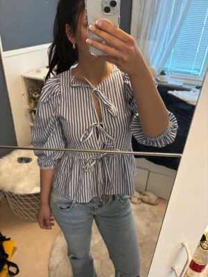 Blårandig blus  - Snygg blus med blåvita vertikala ränder och tre knytningar framtill. Blusen har puffiga axlar och trekvartsärm samt lätt volang nertill. Perfekt för en trendig och avslappnad look. Materialet känns lätt och luftigt.