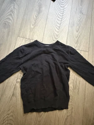 Svart stickad tröja med rund hals - En enkel svart stickad tröja med rund halsringning och långa ärmar. Tröjan har en klassisk passform och ribbstickade muddar vid ärmslut och nederkant. Perfekt att matcha med jeans eller kjol för en stilren look.