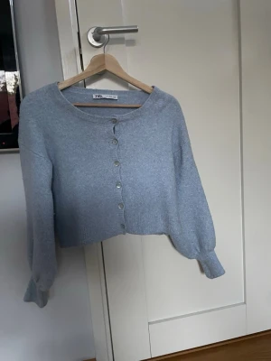 Ljusblå cropped kofta från Zara - Söt ljusblå cropped kofta från Zara med rund halsringning och knappar framtill. Koftan har ballongärmar med breda muddar och är stickad i ett mjukt material. Perfekt att styla över en topp eller klänning.