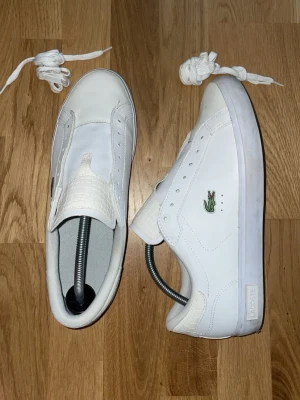Vita Lacoste sneakers  - Snygga vita sneakers från Lacoste med klassisk grön krokodillogo på sidorna. Skorna är använda lite grann men de mesta är smutsigt under skorn. Smörerna följer med vid sidan och kan sättas på själva med egen design. Original box medföljer ej. Pris går att diskuteras vid snabb affär. Perfekta för sommaren/ fin skor.