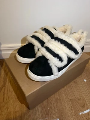 Inuikii - Unika svarta sneakers med breda kardborreband täckta av vit fuskpäls. Skorna har en klassisk låg siluett och vit sula. Perfekta för dig som vill sticka ut med en mysig och trendig look. Materialet är mockaliknande med fluffiga detaljer för extra stil.