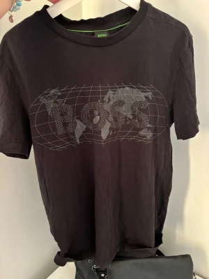 Svart BOSS t-shirt med världskarta - Cool svart t-shirt från BOSS med ett stort tryck av en världskarta och BOSS-loggan på bröstet. Klassisk passform med rund hals och korta ärmar. Perfekt för dig som gillar streetstyle och vill sticka ut lite extra.