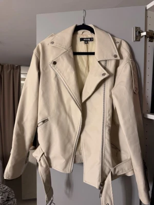Vit skinnjacka från Missguided - Snygg vit skinnjacka från Missguided med klassisk bikerdesign, silvriga dragkedjor och bälte i midjan. Jackan har breda slag, axelklaffar och coola detaljer som ger en edgy look. Perfekt för att lyfta vilken outfit som helst.