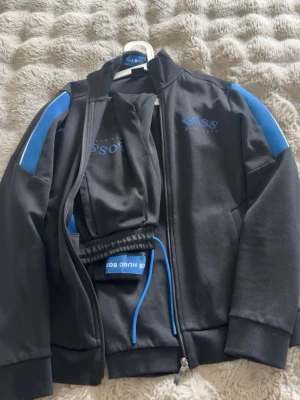 Svart ziptröja från Hugo Boss - Svart ziptröja från Hugo Boss med blå detaljer på axlarna och logga på bröstet. Tröjan har hög krage, dragkedja hela vägen och ribbade muddar. Perfekt för en sportig och stilren look.