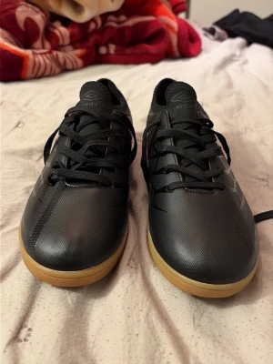 Svarta Umbro fotbollsskor - Snygga svarta fotbollsskor från Umbro med klassisk låg profil och gummisula i naturfärg. Skorna har snörning och är tillverkade i slitstarkt syntetmaterial som ger bra bollkontroll. Perfekta för inomhusspel och futsal.
