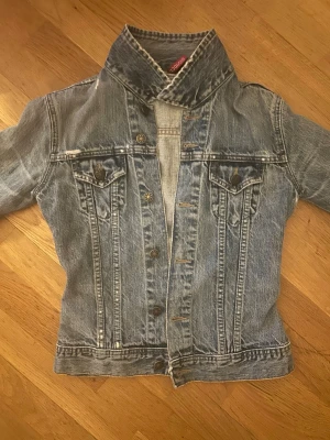 Ljusblå jeansjacka - Snygg blå jeansjacka med klassisk passform och coola detaljer som bröstfickor med lock och knappar framtill. Jackan har hål på ena armen. Jackan har markerade sömmar och justerbara knappar i midjan. Perfekt för dig som gillar en tidlös streetstil.