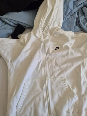 Vit Nike hoodie med dragkedja - Säljer en stilren vit hoodie från Nike med dragkedja framtill och klassisk svart Nike-logga på bröstet. Hoodien har huva med dragsko och två stora fickor framtill. Perfekt för en avslappnad och sportig look priset kan diskutera 