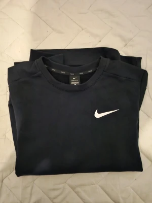 Svart Nike Dri-FIT sweatshirt - Svart sweatshirt från Nike med vit logga på bröstet. Tillverkad i Dri-FIT-material som transporterar bort fukt, perfekt för träning eller chill. Klassisk rund halsringning och enkel design som funkar till det mesta.