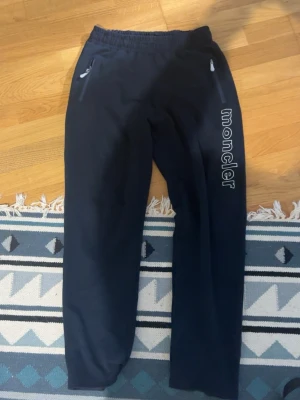 Svarta mjukisbyxor från Moncler - Snygga svarta mjukisbyxor från Moncler med vit broderad logga längs ena benet. Byxorna har resår i midjan och dragkedjeförsedda fickor framtill. Perfekta för en chill och avslappnad stil.