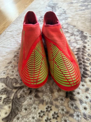 Oranga Adidas fotbollsskor med gröna detaljer - Säljer ett par röda Adidas fotbollsskor med gröna och gula detaljer. Skorna har en strumpliknande passform utan snörning och mönstrad ovandel för bättre bollkontroll. Sulan har gröna dobbar för bra grepp på planen. Perfekta för dig som vill sticka ut på matchen!
