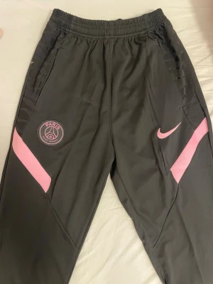 Svarta PSG byxor från Nike, M - Snygga svarta träningsbyxor från Nike med rosa detaljer och PSG-logga på ena benet. Byxorna har elastisk midja med dragsko och två fickor. Perfekta för fotbollsträning eller chill. Materialet är lätt och andas bra.