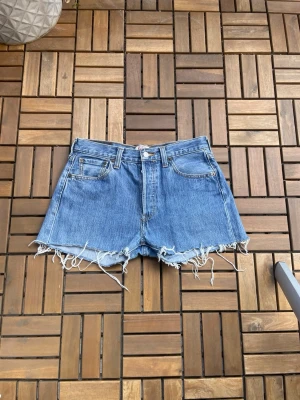 Levi's blå jeansshorts med fransar - Klassiska blå jeansshorts från Levi's med råa, fransiga kanter och femficksdesign. Shortsen har bälteshällor och knappgylf. Perfekta för en avslappnad och trendig look i sommar. Tyvärr för små för mig för att ha baggy, verkligen helt perfekta shorts!!❣️❣️ midjemått 38.5-39