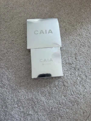 Oöppnad CAIA Soft Focus puder (translusion) och en grå powder puff - Snyggt puder från CAIA i silverfärgad förpackning. Produkten heter Soft Focus och har en stilren, fyrkantig design med spegelblank yta. Den är i färgen translusion. Perfekt för dig som vill ha en smidig basprodukt i necessären. Nypris tillsammans: 490kr! 