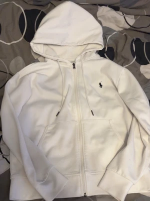 Vit hoodie från Polo Ralph Lauren - Säljer en stilren vit hoodie från Polo Ralph Lauren med dragkedja framtill, huva med dragsko och klassisk logga broderad på bröstet. Hoodien har känguruficka och ribbade muddar vid ärmslut och nederkant. Perfekt för en clean och avslappnad look.