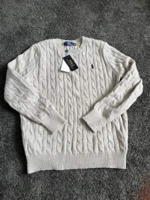 Ljusgrå kabelstickad tröja Polo Ralph Lauren - Snygg ljusgrå kabelstickad tröja från Polo Ralph Lauren med klassisk rund halsringning och svart broderad logga på bröstet. Tröjan har ribbade muddar vid ärmslut och nederkant. Perfekt för dig som gillar stilrena och tidlösa plagg.