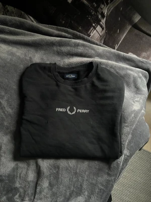 Svart Fred Perry sweatshirt - Svart sweatshirt från Fred Perry med vit broderad logga och text på bröstet. Klassisk rund halsringning och ribbade muddar vid ärmslut och midja. Få skavanker som ett litet mini hål s som passar M