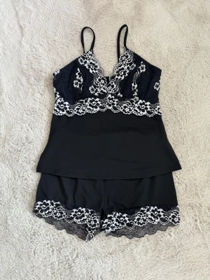 Svart spets-set med linne och shorts - Snyggt svart pyjamasset. Setet har tunna axelband och detaljer i vit spets både på linnet och shortsen. Materialet är mjukt och stretchigt. 