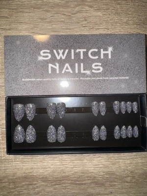 Switch Nails Shadow Flash press-on - Snygga press-on naglar från Switch Nails i modellen Shadow Flash. Naglarna har en kort mandelform och är täckta av silverglittrigt material. Tillverkade av återvunnet material och kan återanvändas. Perfekt för dig som vill ha salongskänsla snabbt. Säljes utan tillbehör.