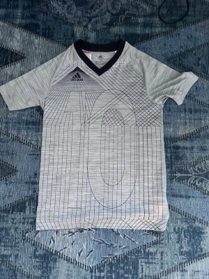 Grå Messi sporttröja Adidas med nummer 10 - Snygg grå sporttröja från Messi och Adidas med svart V-ringning och stort grafiskt nummer 10 på framsidan. Tillverkad i ett lätt och andningsbart material, perfekt för träning eller fotboll. Diskret Adidas-logga på bröstet.