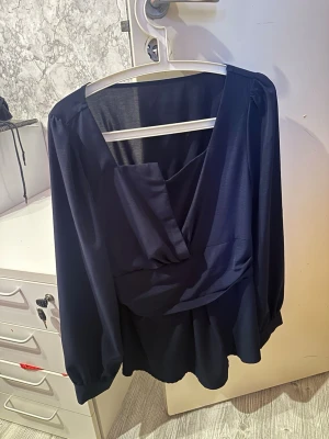 Mörkblå omlottblus med v-ringning - Snygg mörkblå omlottblus med djup v-ringning och smockad midja baktill. Blusen har långa ärmar med lätt puff och är tillverkad i ett tunt, lätt material som ger ett luftigt intryck. 