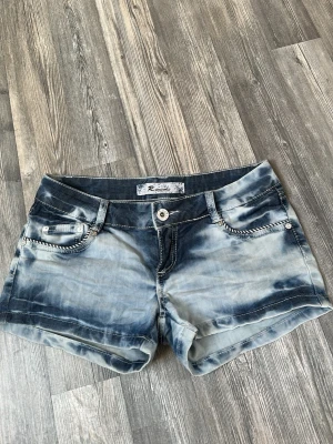 Blå tie-dye shorts från Reistyle - Snygga blå tie-dye shorts från Reistyle med coola detaljer på fickorna och knappstängning. Shortsen har låg midja och är gjorda i bomull, perfekta för en avslappnad stil. Dekorativa sömmar och mönstrade detaljer på bakfickorna ger extra edge.