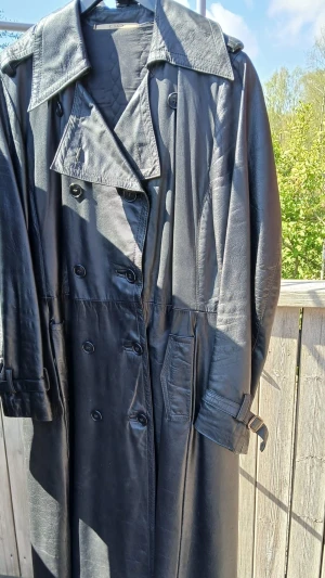 Svart lång skinn trenchcoat för herrar - Snygg svart lång skinn trenchcoat med dubbelknäppning, bred krage och bälte i midjan. Jackan har fickor på sidorna och justerbara spännen vid ärmsluten. Perfekt för dig som vill sticka ut med en klassisk och cool look.