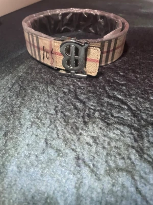 Beige rutigt bälte från Burberry - Stilrent bälte från Burberry i klassiskt beige rutigt mönster med silverfärgat B-spänne. Perfekt accessoar för att lyfta din outfit med en ikonisk touch. Tillverkat i skinn med detaljer i metall.