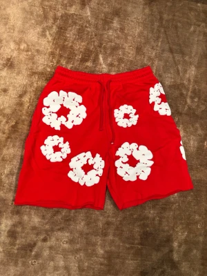 Röda Denim Tears Shorts M - Snygga röda shorts med stora vita blommönster och elastisk midja med snörning. Perfekta för sommaren och ger en riktigt chill vibe. Lätta och bekväma att bära, passar till både strand och häng med vänner. Storlek M
