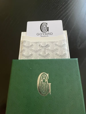 Vit kortfodral från Go4yard - Snyggt vitt kortfodral från Go4yard med klassiskt mönster och präglad logotyp framtill. Tillverkat i canvas och läder, har flera fack för kort och levereras med original grön ask. Perfekt för dig som vill ha en stilren och exklusiv accessoar.
