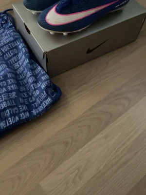 Nike mercurial vapor 16 strl 43 (ag elite)  - Säljer ett par blå Nike fotbollsskor med vita och rosa detaljer. Skorna har klassisk design med dobbar för gräsplan och är tillverkade i syntetmaterial. Liten defekt finns vid strumpan. Medföljer en blå Nike-påse med tryck och originalkartong. Perfekt för dig som vill ha stil på planen.