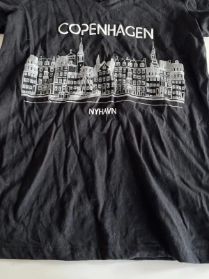 Svart t-shirt Copenhagen print - Svart t-shirt med vit tryckt illustration av Nyhavn i Köpenhamn och texten 'Copenhagen' på bröstet. T-shirten har v-ringning och korta ärmar, perfekt för en avslappnad streetstyle-look.