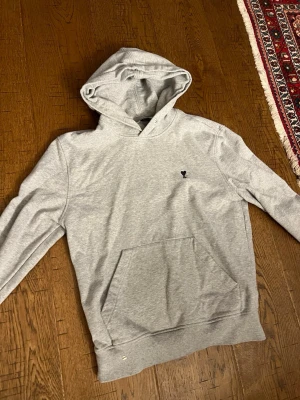 Ami Paris hoodie  - Snygg grå hoodie från AMI Paris med svart hjärtdetalj broderad på bröstet. Klassisk känguruficka framtill och justerbar huva. Tillverkad i mjuk bomullsblandning som känns skön mot huden. Perfekt för en avslappnad och trendig look.