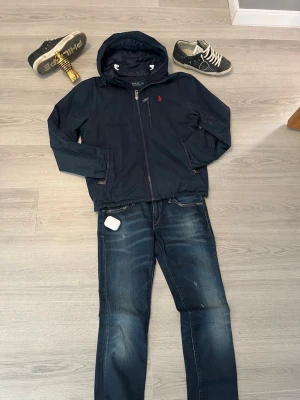 Marinblå vindjacka Polo Ralph Lauren - Snygg marinblå vindjacka från Polo Ralph Lauren med huva och dragkedja framtill. Jackan har en liten röd logga på bröstet, praktiska fickor och justerbara muddar. 
