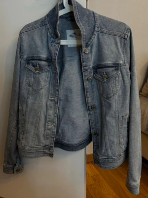 Ljusblå jeansjacka från Hollister - Snygg ljusblå jeansjacka från Hollister med klassisk design, bröstfickor med lock och knappar samt raka sömmar. Jackan har en stilren krage och silverfärgade knappar framtill. Perfekt för dig som gillar en avslappnad och trendig look.