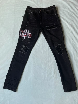 Svarta ripped jeans med brodyr - Snygga svarta jeans med slitningar och broderad patch i rött och vitt på låret. Jeansen har skinny fit och klassisk femficksdesign. Perfekta för en edgy streetstil.
