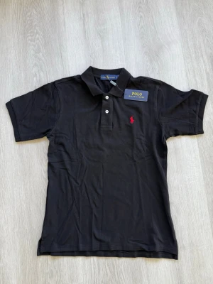 Svart pikétröja från Ralph Lauren - Klassisk svart pikétröja från Ralph Lauren med röd broderad logga på bröstet. Tröjan har krage, korta ärmar och knäppning med tre knappar framtill. Perfekt för en stilren och avslappnad look.