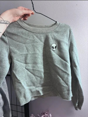 Grå sweatshirt med alien-tryck - Ganska liten i storleken, passar Small. Grå sweatshirt med rund halsringning och ett litet broderat alien-märke på bröstet. Tröjan har långa ärmar och är gjord i mjukt sweatshirtmaterial, perfekt för en avslappnad stil.